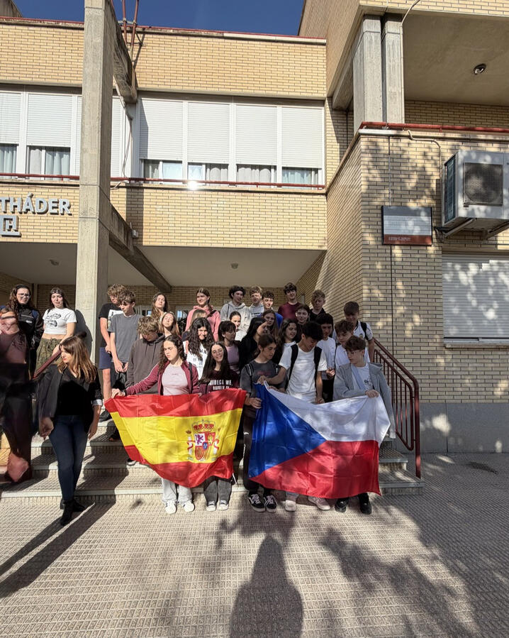 Společná fotografie českých a španělských studentů před španělskou střední školou. Studenti drží vlajky obou zemí jako symbol mezinárodního přátelství a spolupráce v rámci výměnného programu. Čeští a španělští středoškoláci společně drží české a španělské vlajky před španělskou školou